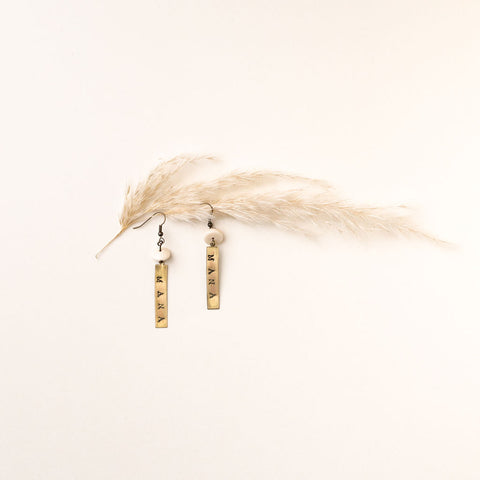 A pair of the Mana earrings from Koakoa Design. In the image the earrings are laid flat on a pale warm cream background. They look as if they're hanging from a small toetoe frond. The earrings have brass plaques on which the word "MANA" is stamped. The plaques are affixed to small rounded bone buttons which are in turn joined to earring hooks.
