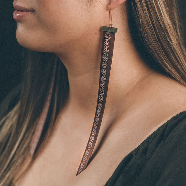 A close up of the Kia Māia earrings worn on a model. The image shows the partial face and shoulders of the model who is wearing a dark green linen top. She has long brown hair with golden highlights. The earring shown, is draped against her neck and ends at her collar bone. The earring is in toffee coloured satin ribbon with a mangopare printed designed in white.