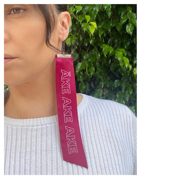 Partial profile of a Māori woman wearing a red satin earring with Āke Ake Ake printed in capital letters.