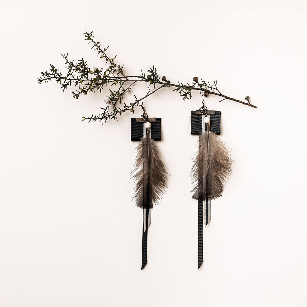 Styled product image of the Amaru earrings by Koakoa Design. The earrings feature black and cream ribbons, on top of which lay a chain topped with a cream Howlite stone and a black feather.  The earrings are placed as if hanging from a Mānuka twig with a small number of white flowers.
