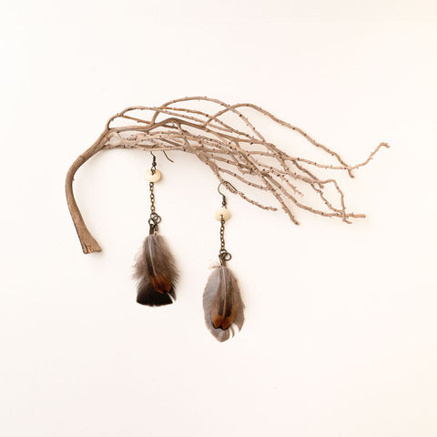 A pair of the Koakoa Design Ngā Manu earrings lie on a pale cream background. The look as if they're hanging from a small branch. The earrings have small bone beads from which two feathers dangle from a chain.