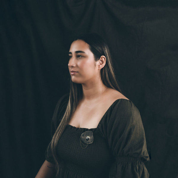 Image showing a young Māori woman in a dark olive dress wearing the Anchored brooch by Koakoa Design. The model has long hair and is sitting in front of a dark linen backdrop. The image is moody.