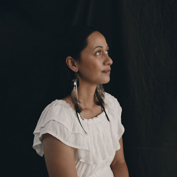 A model is wearing the Amaru earrings. She is a Young Māori woman who is facing to the right of the shot. Her face an upper torso show in profile. She is wearing a white frilled short sleeved short and her black hair is pulled back from her face. She sits in front of a black linen backdrop and the shot is moody.