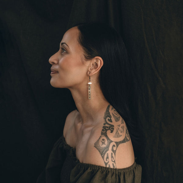 A young Māori woman wears the Mana earrings from Koakoa Design. She is in an off the shoulder dark olive top. She is facing the left and her face and upper torso are in profile. She has a Māori tattoo visible on her back and her long black hair is tucked behind her ears and flowing loose down her back. She has a small smile on her face.