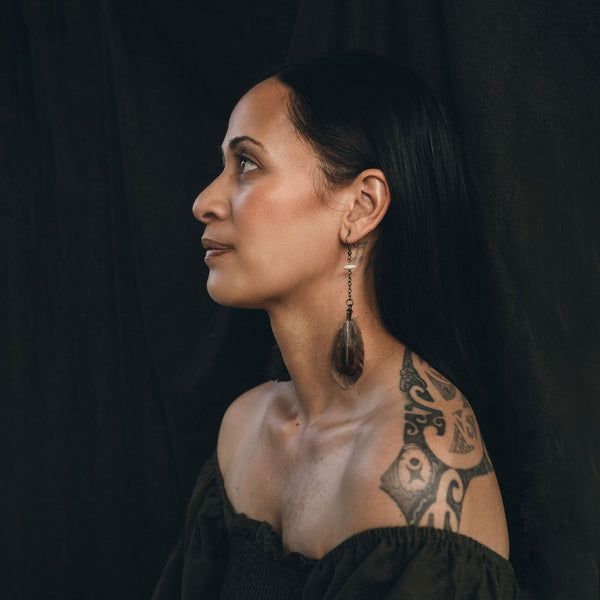 A Māori woman is adorned with Ngā Manu earrings. She looks to the left of the shot and her face and upper torso are in profile. Her Māori designed tattoo is visible on her back and she wears a dark green off the shoulder top. Her long dark hair is tucked behind her ear and flows down her back. She looks thoughtful and is pictured against a dark background. The image is moody.