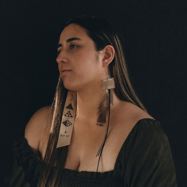 A young Māori woman wears the Ko Au earrings from Koakoa Design. She is looking slightly to the left of the shot and looks sombre. Her hair is dark shot with golden highlights and she wears an off the shoulder dark olive top. She sits in front of a dark black background and the shot is moody. The Māori design earrings drape against her neck and chest.
