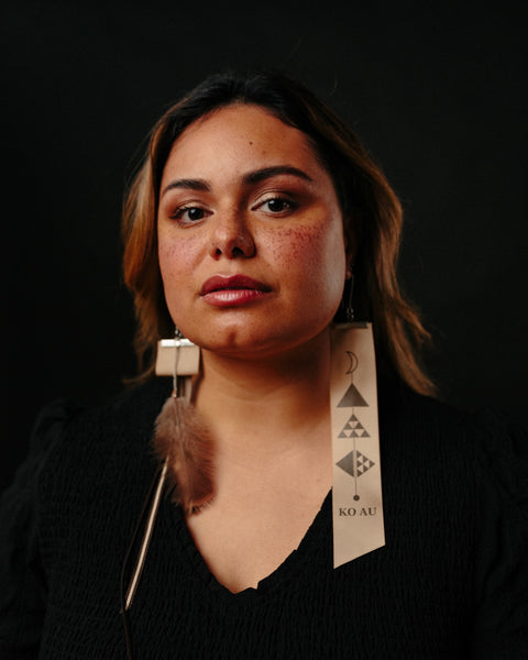 A Māori woman wears the Koakoa Design Ko Au earrings. Her caramel brown hair flows down her shoulders and she is wearing a v neck black top. Her head is tilted up slightly and she gazes down towards the camera. One earring has a graphic representing pēpeha, the other has a korukoru feather.