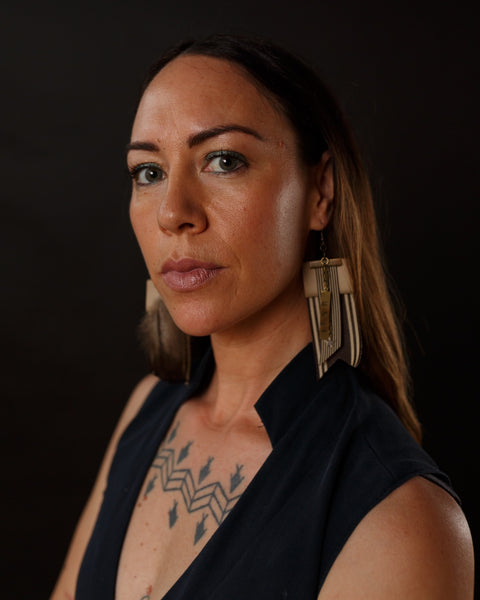 A woman is wearing the Haki earrings from Koakoa Design. She has a sideways stance but has turned her face towards the camera. She has a v-neck black top which shows the tattoos just under her collar bone. Her gaze is steady but she has an air of pensiveness about her.