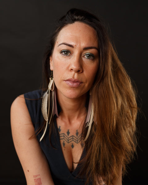A brown skinned woman is wearing the Old Bones earrings from Koakoa Design. She looks straight into the camera. Her hair falls to one side of her face and her short sleeved black v neck top shows parts of the tattoos on her arm and below her collar bone.