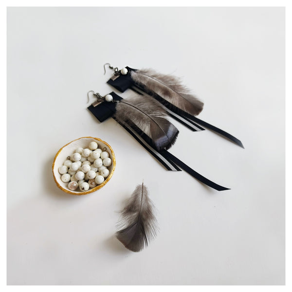 A pair of Amaru earrings laying flat on a pale grey background. To the foreground of the image is a single black feather which also adorn each earring. To its left is a small cream clay dish with gold edging inside of which are a pile of cream Howlite stones.