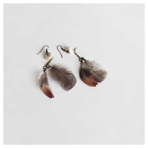 A pair of earrings lie on a pale background. Each earring has two small natural feathers in browns, greys and black which are attached to an antiqued chain which connects to a rounded bone bead which joins to a shepherd hook style earring hook.